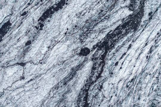 Aerial view of a glacier featuring intricate patterns of blue ice and dark volcanic ash deposits across the frozen landscape in ICELAND.
