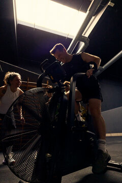 Young man and friends pushing through an intense air bike workout in a dimly lit gym