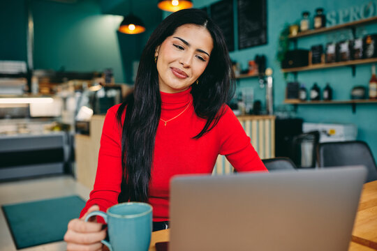 Smiling woman in red sweater using laptop at a coffee shop with mug for remote work and productivity