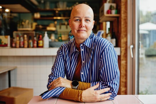 Portrait of a bald person in a blue striped shirt sitting in a cafe, looking at the camera