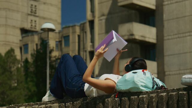 A young high school student relaxes outdoors, engrossed in her notebook while listening to music through headphones. She enjoys a peaceful study session outside on a bright, sunny day.
