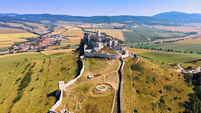 Spis Castle fortification walls and towers seen from above. High aerial shot of medieval stone walls extending along green ridge toward main castle keep, bright daylight.