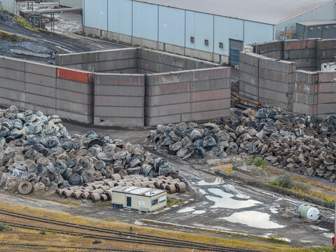 Aerial view of Tata Steel industrial site featuring large piles of scrap metal, concrete storage walls, and a small office building in Velsen-Noord, Noord-Holland, Netherlands.