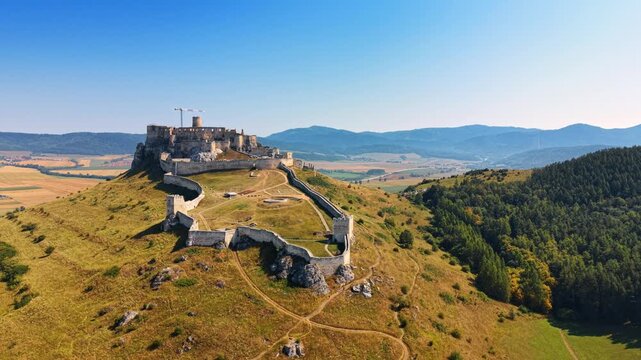 Castle sits on hilltop in sunny weather. A castle stands on a hill surrounded by fields and trees in the distance. The sky is clear and blue.