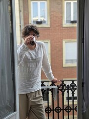 Fototapeta premium Man in a white linen shirt drinking coffee on a balcony on a calm summer morning