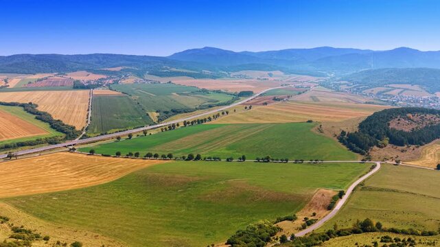 Aerial view of patchwork agricultural fields and roads. Green and yellow farmlands stretch toward distant mountains under a bright blue sky.