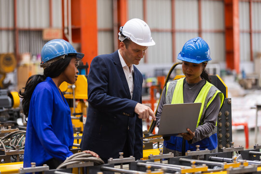 Manufacturing team reviews production data on laptop beside industrial machinery in a modern factory, representing engineering, quality control, teamwork, safety, digital monitoring.