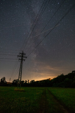 Electric power lines and pylon stretching across a green field under a clear starry night with the Milky Way.