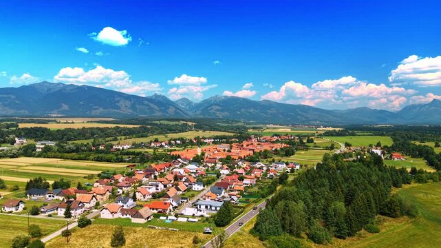 Panoramic view of rural village near High Tatras mountains. Aerial view of village surrounded by forest, fields, and mountain range under blue sky with clouds.