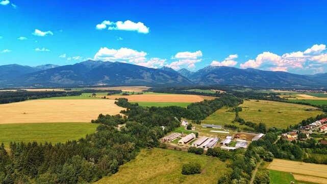 Agricultural farm complex against High Tatras mountain range. Aerial view of farm buildings, farmland, and green trees with mountain peaks in background under blue sky.