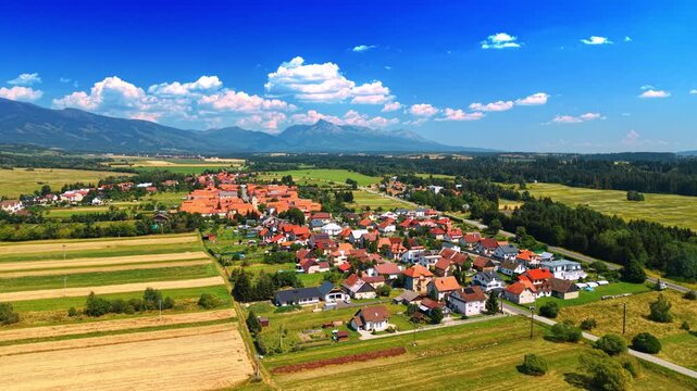 Slovakian village with red roofs near High Tatras mountains. Drone view of rural houses and roads bordered by farm fields and mountain peaks under cloudy sky.