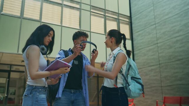 High school students share headphones and notes outside a modern school building. Friends enjoy a lighthearted interaction, connecting over music and studies on campus.