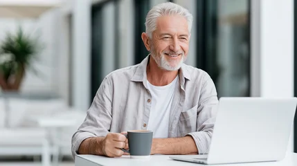 Senior man with gray hair smiles while holding a coffee cup and using a laptop at a bright indoor workspace with plants in the background © Nataliia