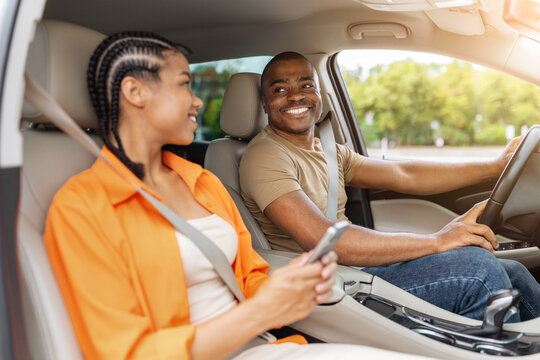 Young Black couple smiling at each other inside a modern car, with the man at the wheel and the woman holding smartphone, depicting connected road travel and lifestyle