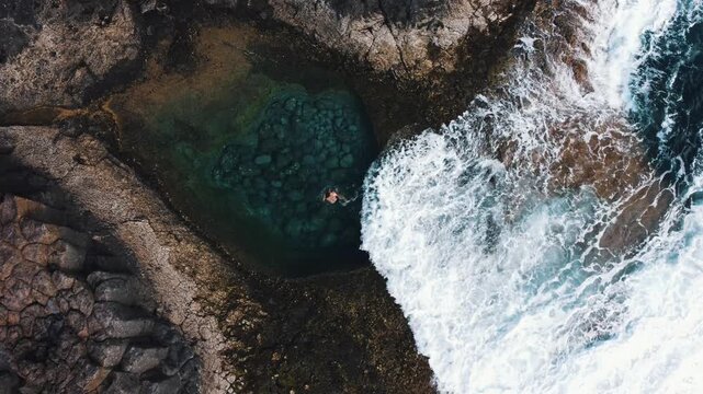Aerial view of a woman relaxing in a natural tidal pool as ocean waves crash against the rocks