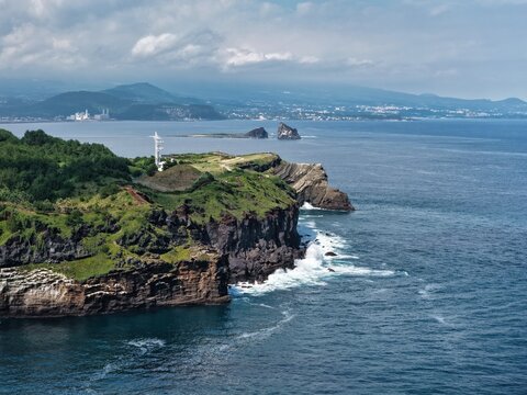 Aerial view of the rugged coastal cliffs of Jeju Island with green vegetation, a white lighthouse, and crashing waves in the blue sea, South Korea.
