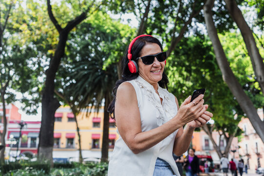 senior latina woman using headphones and listening music in public park in Mexico Latin America, middle age female