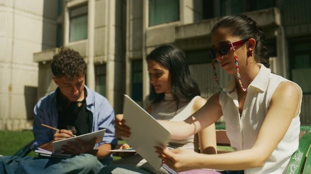 Three high school students are sitting on a park bench, engaging in study and discussion outdoors. This group of young people is focused on their learning activities together in a casual setting.