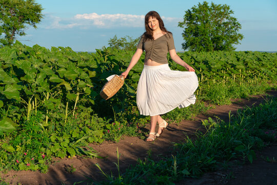 Young woman in white skirt walking through sunflower field with wicker basket