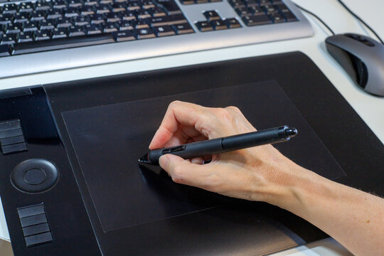 Designer's hand draws with a digital stylus on a black graphics tablet on a desktop next to a computer keyboard