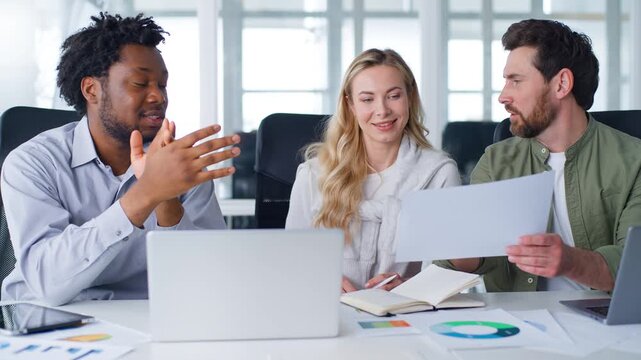 Three smiling professionals multiracial business colleagues talking about papers data information discussing trading startup report at boardroom office table meeting team managers talk with document