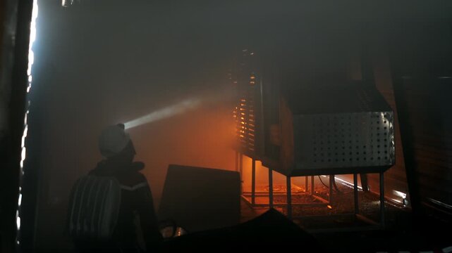 Firefighter wearing protective gear inspects a smoke filled fire scene using a headlamp. Emergency responder assessing a hazardous industrial area after a fire.