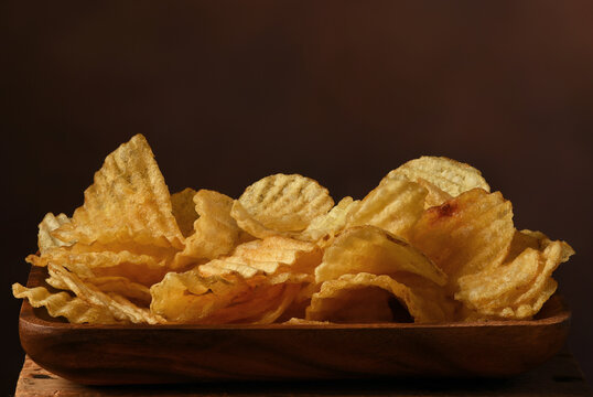 Still life of a wood bowl filled with Potato Chips againsa a waem brown mottled background.