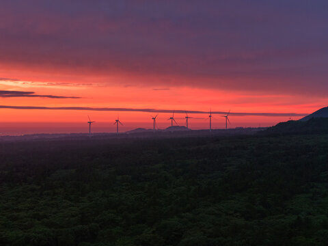 Aerial view of wind turbines in a wind farm at sunset with a vibrant red and orange sky over a forested landscape in South Korea.