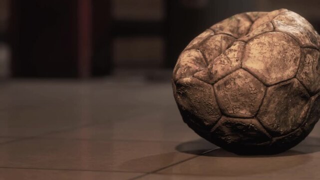 A worn soccer ball rests on the floor in a dimly lit room. The surface of the ball shows signs of use and age. Shadows are visible in the background, creating an atmosphere of simplicity.