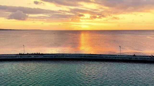Aerial view of the Marina Togo Wakatobi pier with people sitting along the structure during a golden sunset over the sea in Kabupaten Wakatobi, Sulawesi Tenggara, Indonesia.