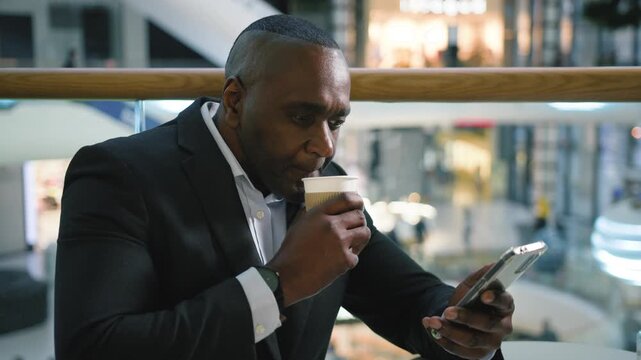 Relaxed middle-aged African American business man drinking coffee chatting mobile phone network in cafe mature businessman male user drink tea cappuccino using smartphone device in cafeteria break