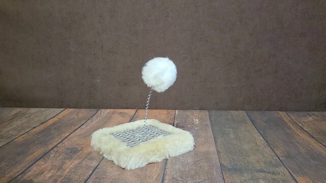 White fluffy pompom on a metal spring attached to a plush cat scratching pad swings on a wooden floor against a brown background. Close up of a pet toy for cats and kittens.