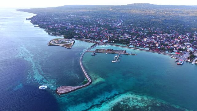 Aerial view of Marina Togo Wakatobi with its curved pier extending into the clear turquoise sea, adjacent to the coastal town and lush greenery in Kabupaten Wakatobi, Sulawesi Tenggara, Indonesia.