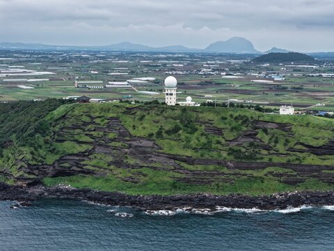 Aerial view of the radar station atop the coastal cliffs of Suwolbong Peak on Jeju Island, surrounded by lush green vegetation and agricultural fields in South Korea.