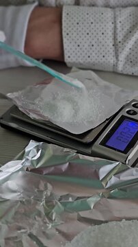 A chemist pours white powder into a small plastic bag on a digital scale in a lab. A doctor fills a mailing bag with drugs in a crime lab.