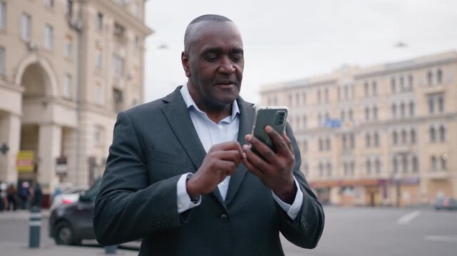 Smiling African American 50s mature business man talking mobile phone video call walking in city outdoors confident middle-aged businessman male executive talk conference with smartphone chat app walk