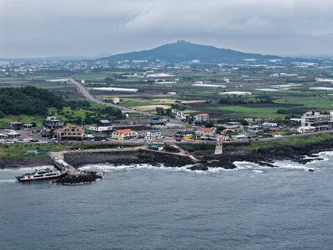 Aerial view of a coastal town with a ferry docked at a pier, surrounded by farmland and a volcanic cone in the background in South Korea.