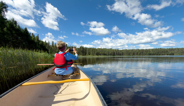 Senior woman sitting in the front of a canoe talking a picture of a calm lake and blue summer sky
