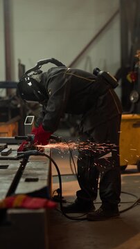 Vertical video of industrial worker in protective gear using angle grinder to cut metal, creating bright sparks. Footage is suitable for manufacturing promos or safety training videos
