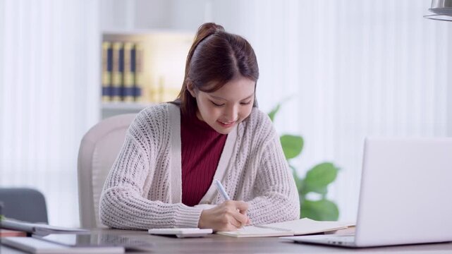 Young Asian woman writing notes in notebook while studying or working at modern home office desk