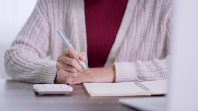Young Asian woman writing notes in notebook while studying at home desk with calculator and copy space