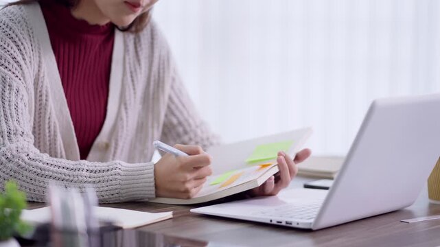 Young Asian Muslim woman writing notes from laptop during online learning session at home office desk