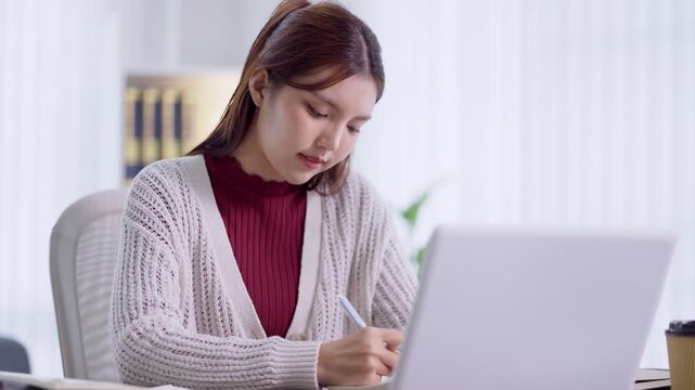 Young Asian woman writing notes while studying on laptop in bright home office with copy space