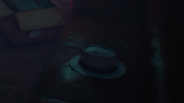 A hat is placed on a leather seat in a dimly lit room. Soft shadows surround the hat, and a few objects are visible in the background. The atmosphere feels intimate and quiet.