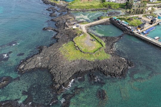 Aerial view of the volcanic rock coastline with clear turquoise water, a small harbor, and a grassy patch in South Korea.
