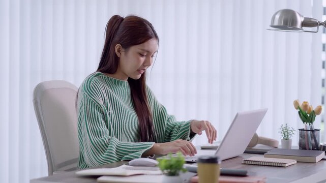 Young Asian woman typing on laptop at modern home office desk in bright natural light with copy space