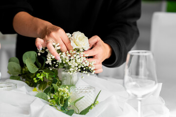 Fototapeta premium A woman's hands arranging delicate white baby's breath and a rose in a small glass vase on a decorated wedding table.