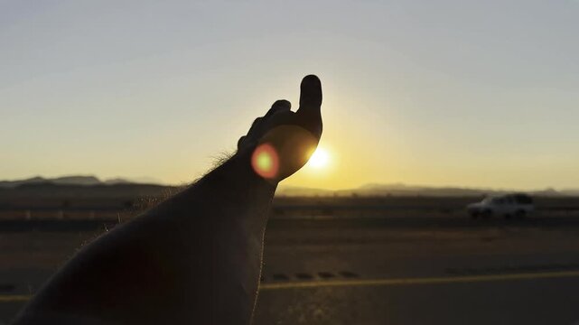 Silhouette of person arm reaching towards sun on desert highway during sunset
