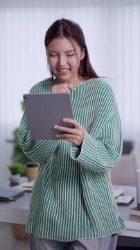 Young Asian woman smiling while using digital tablet in modern bright home office workspace