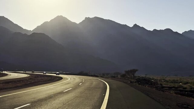 Scenic mountain highway with cars driving under bright sunbeams behind mountain peaks